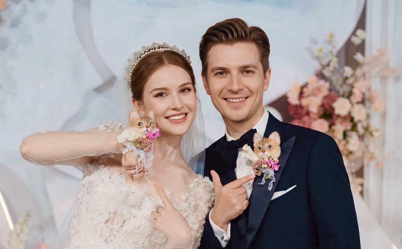 A smiling bride and groom in wedding attire holding custom 3D pet portrait brooches, featuring personalized handmade wool felt cat wedding boutonnieres on their lapels.