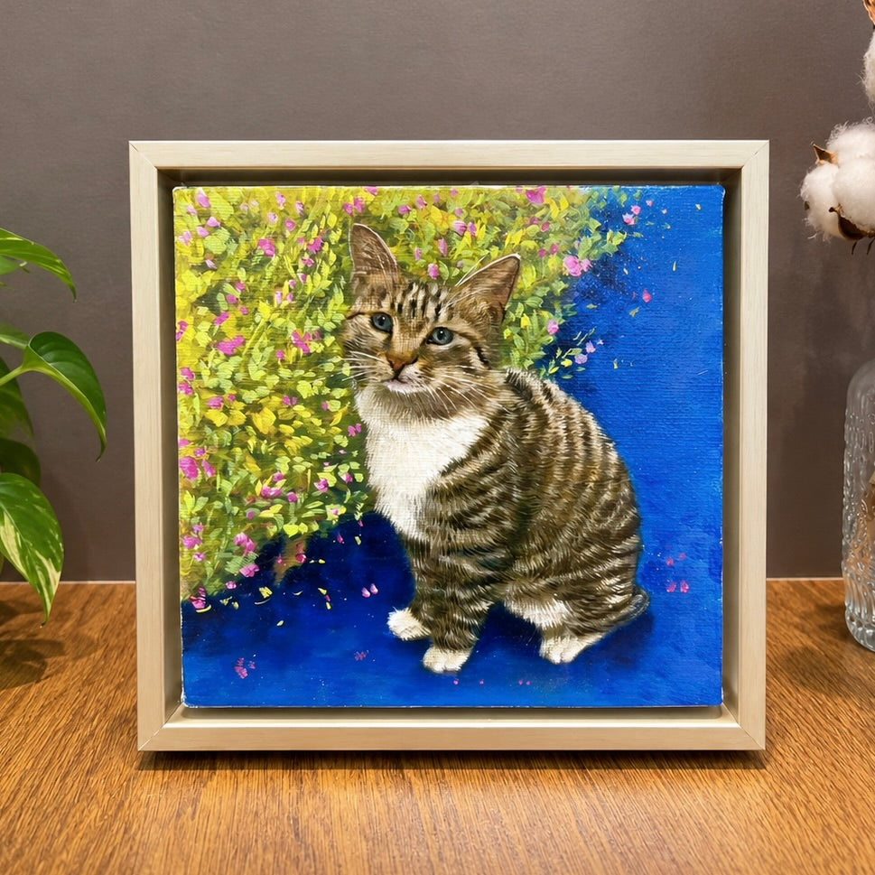 Custom hand-painted oil portrait of a tabby cat with white fur on its chest, presented in a light wooden frame next to dried cotton flowers.