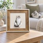 Brown framed 3D portrait of a seated silver-gray cat on a table, with a cat playing on the sofa behind it