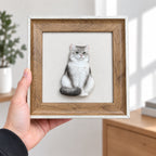Hand holding a brown frame with light outer trim and a full-body silver-gray cat portrait with its tail curled forward