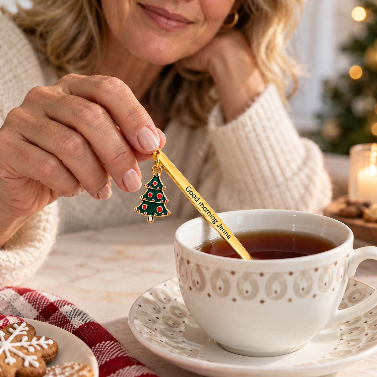 A gold-tone personalized Christmas spoon with a Christmas tree charm resting beside a cup of tea on a festive morning.