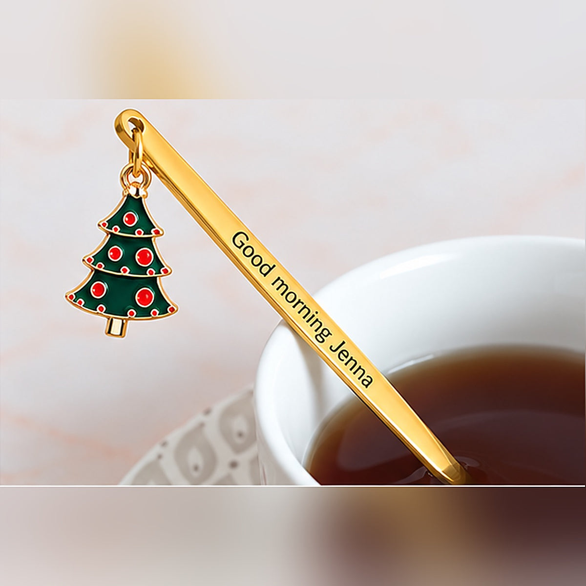 Gold-tone personalized spoon with custom text and a green Christmas tree charm beside a cup of tea