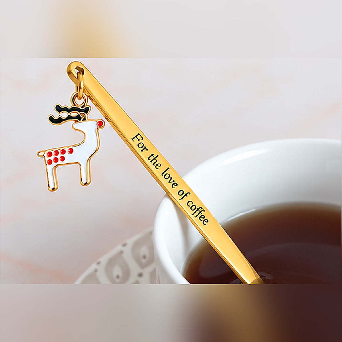 Gold-tone personalized spoon with custom text and a white reindeer charm beside a cup of tea