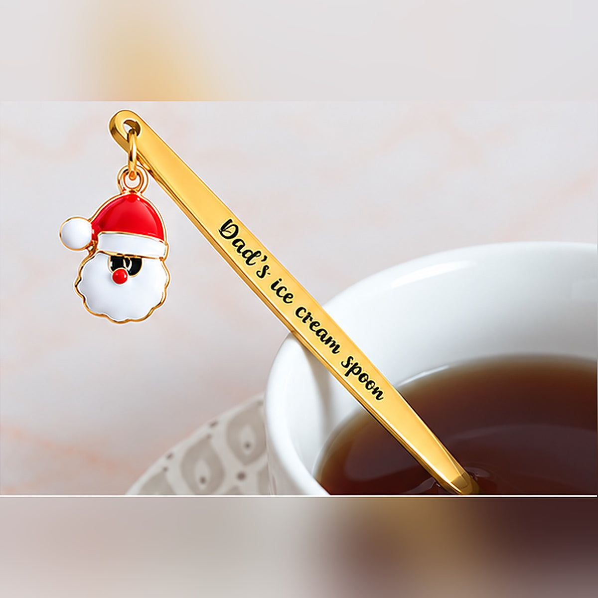 Gold-tone personalized spoon with custom text and a Santa Claus charm beside a cup of tea