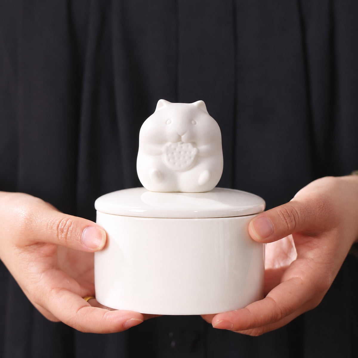 White ceramic hamster memorial urn held in two hands against a dark background