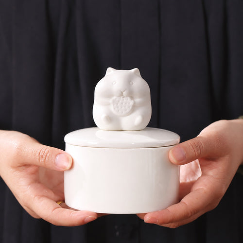 White ceramic hamster memorial urn held in two hands against a dark background