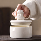 Hand lifting the hamster-shaped lid from a white ceramic memorial urn to show the interior opening