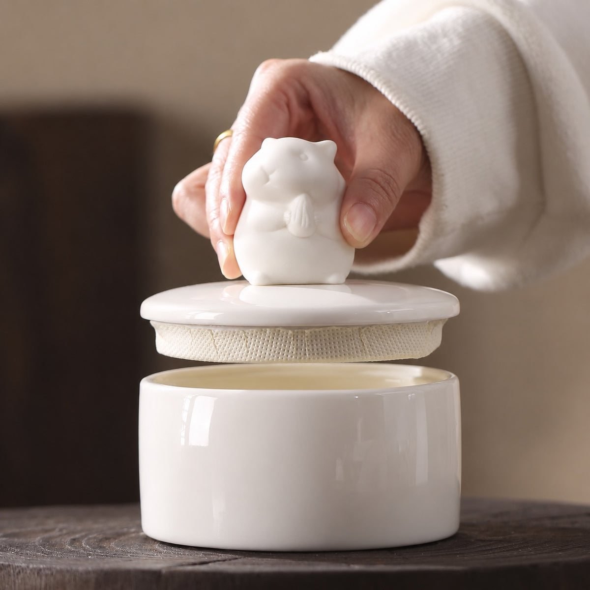 Hand lifting the hamster-shaped lid from a white ceramic memorial urn to show the interior opening