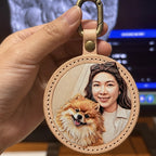 A hand holds a custom, round leather keychain with a hand-painted 3D relief carving of a woman and her pomeranian dog, featuring stitched edges and a gold clasp.