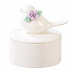 Ivory ceramic bird memorial urn with a removable lid topped by a bird figure, purple flower and green leaves