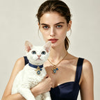 A woman holding a white cat, both wearing a matching set of custom shared human-pet jewelry. The owner wears a gold necklace and the cat wears a blue collar, each featuring a blue birthstone and photo locket charm.