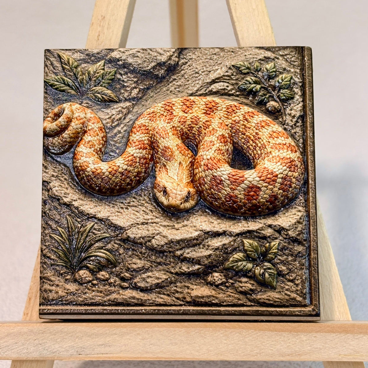 Orange patterned snake relief artwork with textured rocks and leaves, displayed unframed on an easel