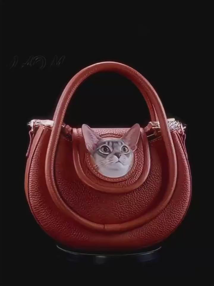 A uniquely shaped, handcrafted brown textured pebble-grain leather handbag featuring a circular cutout with a highly detailed 3D leather carving of a grey and white shorthair cat looking upward. Designed and handcrafted by a luxury bespoke leather artisan workshop in memopaw.