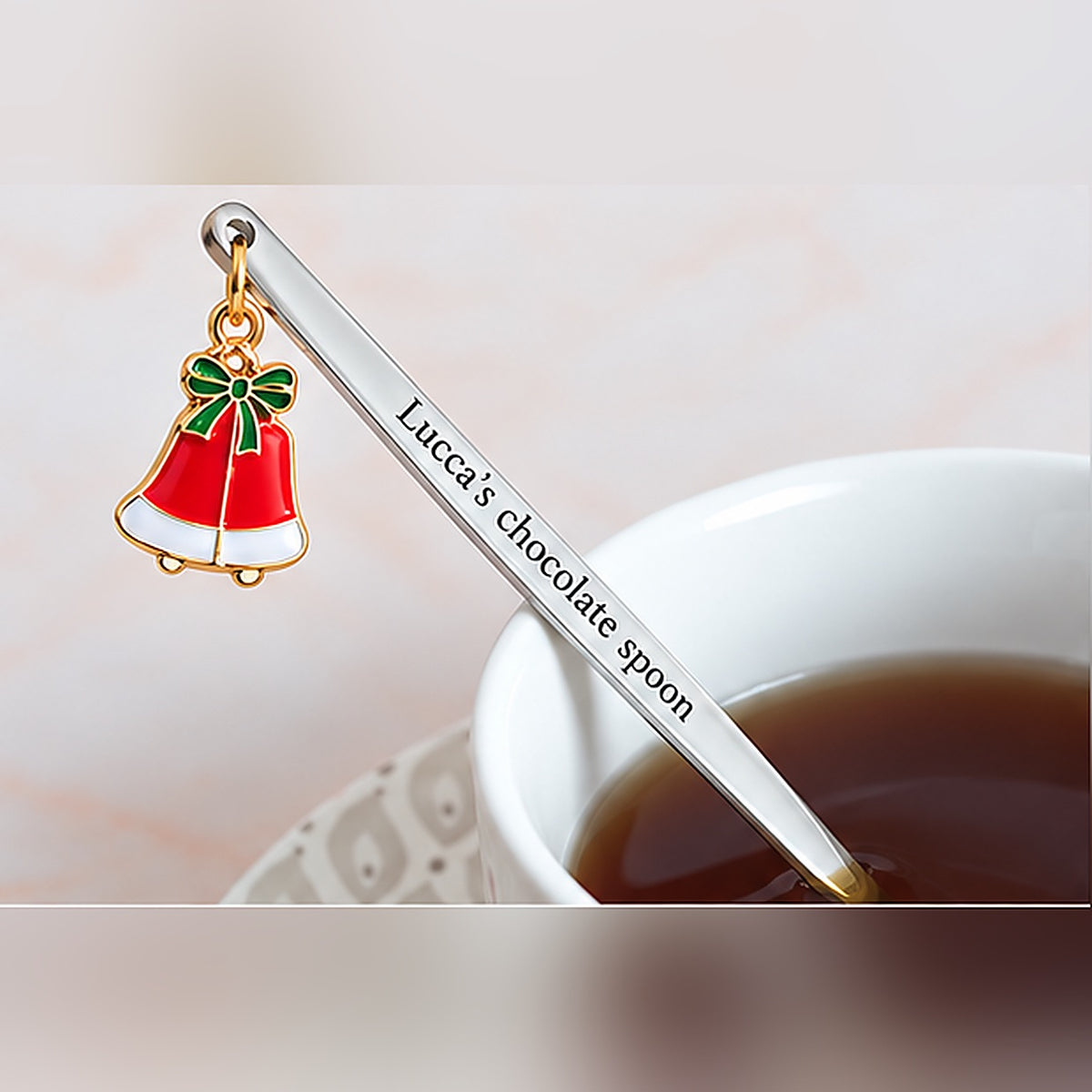 Silver-tone personalized spoon with custom text and a red Christmas bells charm beside a cup of tea