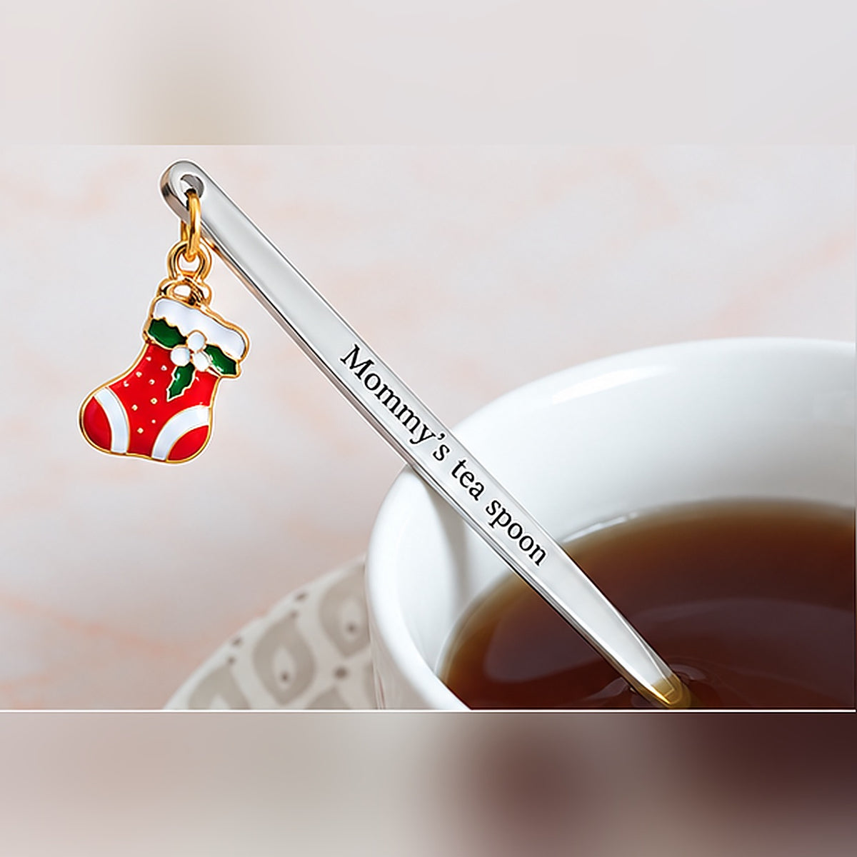 Silver-tone personalized spoon with custom text and a red Christmas stocking charm beside a cup of tea