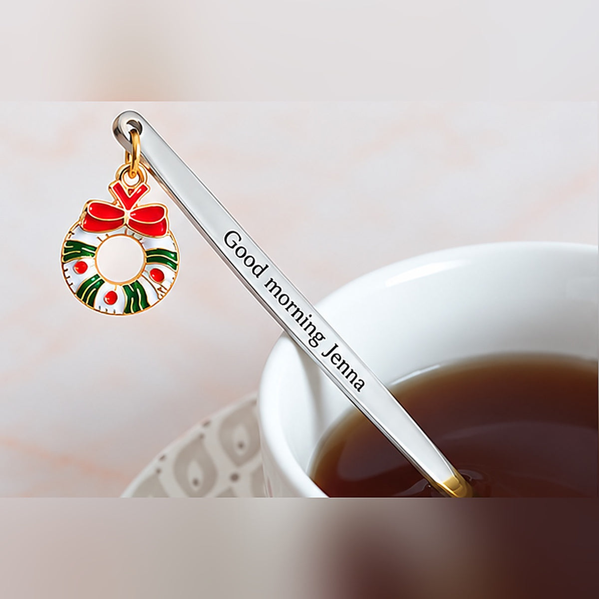 Silver-tone personalized spoon with custom text and a red and green Christmas wreath charm beside a cup of tea