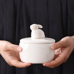 White ceramic sitting rabbit memorial urn held carefully in two hands against a dark background