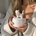 Person holding a small ivory ceramic cat memorial urn with pink flower and green leaf decoration