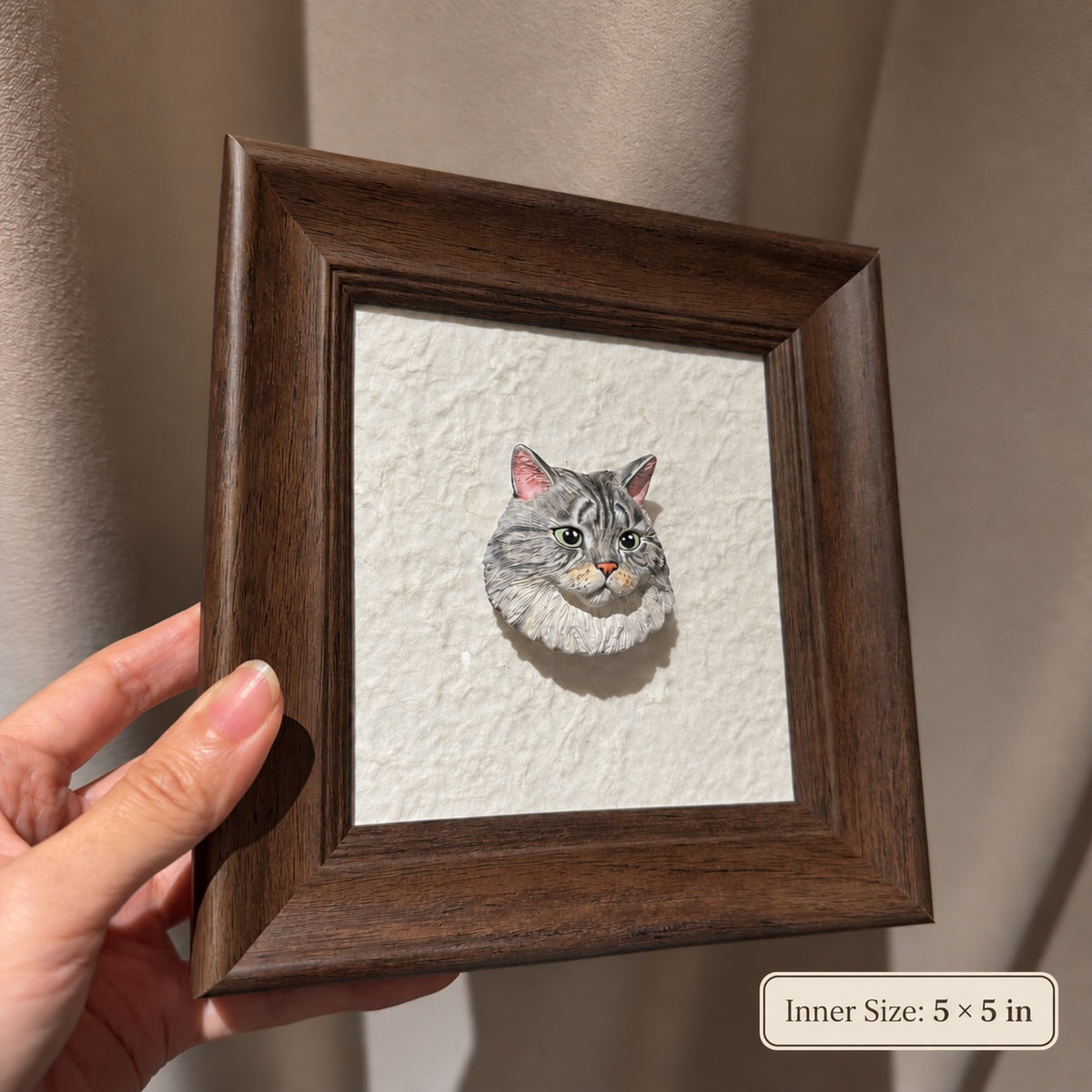 Gray tabby cat head portrait in a dark brown frame, held by a hand and labeled 5 by 5 inches inner size