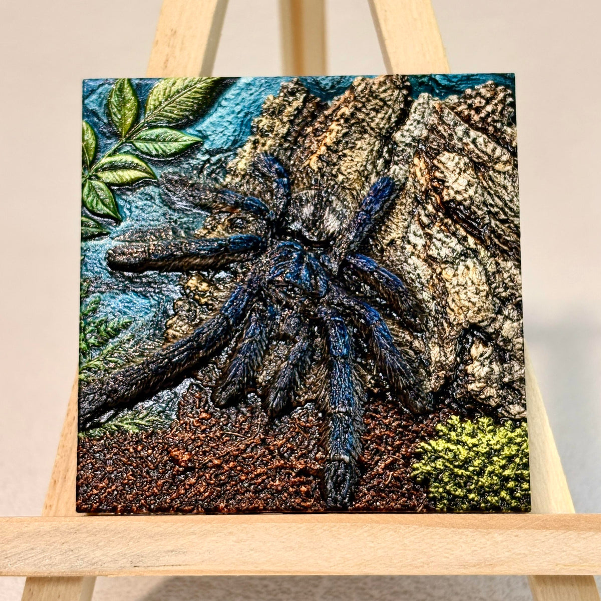 Unframed relief artwork of a blue-toned tarantula against textured bark and leaves on an easel