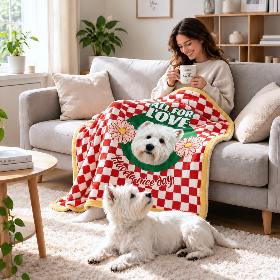 Person holding a mug under a red dog portrait blanket with a white dog sitting beside the sofa